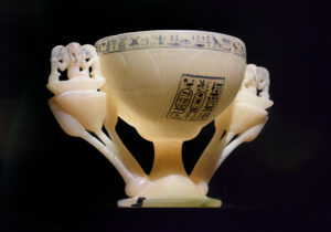 The lotus chalice is carved from a single piece of alabaster. The chalice takes the shape of a white lotus in full bloom, identified by its rounded petals. The supports for the handles are shaped like blue lotus flowers which are flanked by buds growing upward, with the god Heh seated on a basket (the neb symbol) on the tips of the petals.